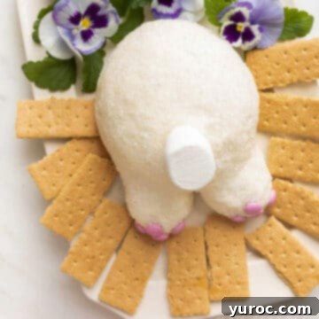 A whimsical bunny butt cheeseball adorning a pristine white platter, accompanied by graham crackers and delicate pansy flowers in the background.