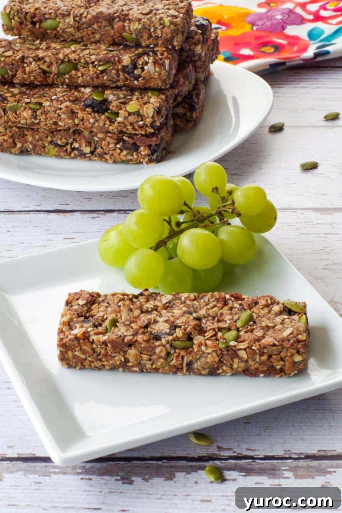 A single No Bake Almond Butter Granola Bar on a pristine white plate, accompanied by fresh green grapes. In the soft-focus background, a platter holds several more delicious granola bars.