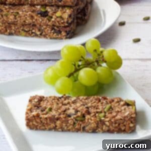 A single almond butter granola bar on a white plate, garnished with green grapes, with a pile of similar granola bars in the background.