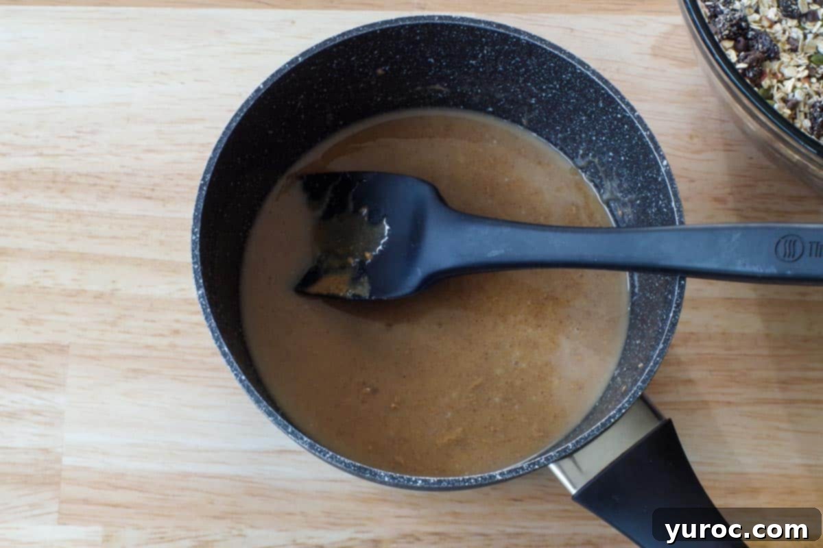 A smooth, melted mixture of coconut oil, almond butter, and honey gently simmering in a saucepan, being stirred with a black silicone spatula.