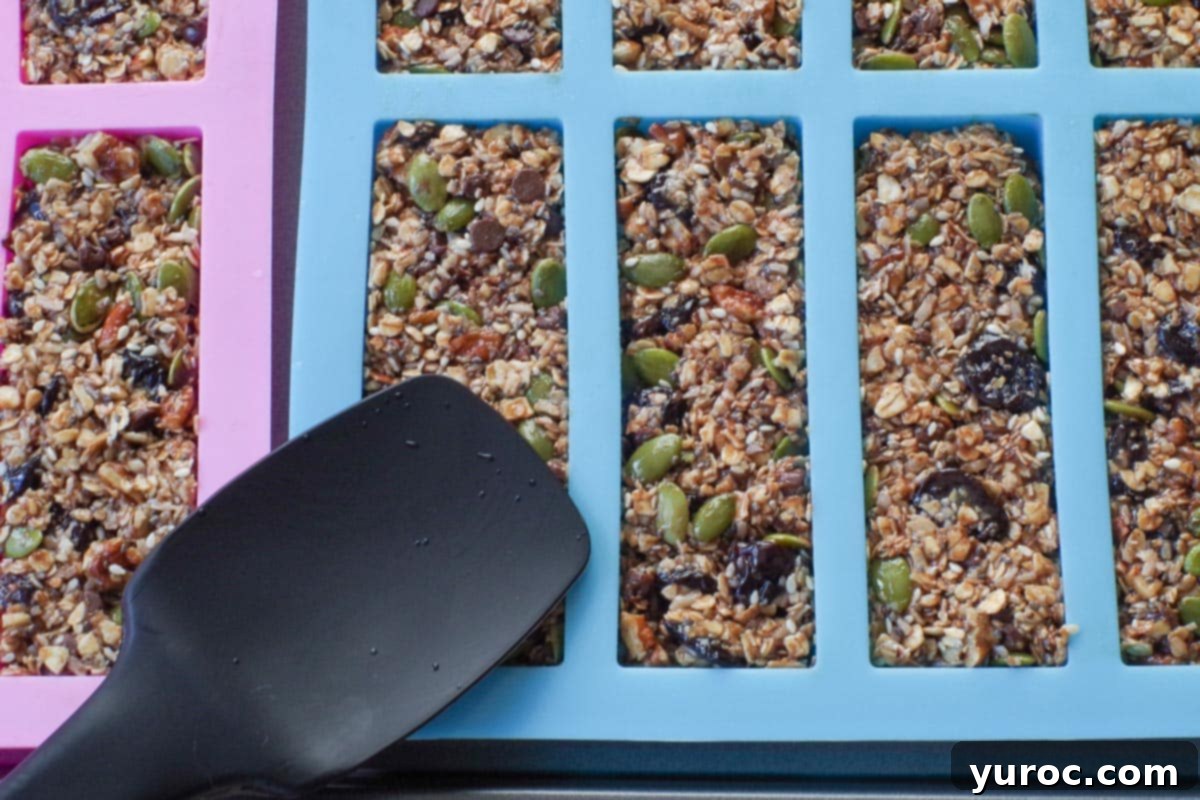 A black spatula is firmly pressing the granola mixture into individual bar molds, ensuring a compact and even surface.