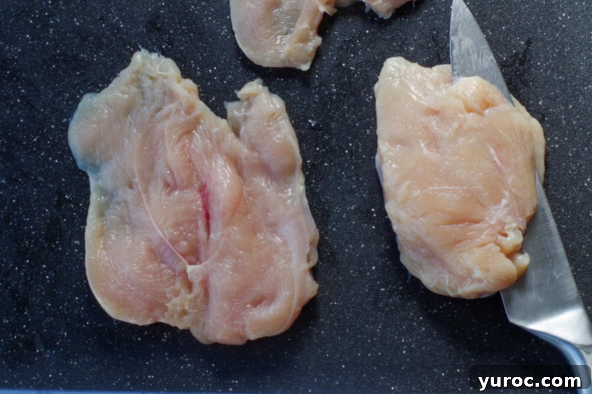 chicken being sliced with a knife on the cutting board, with another piece of raw chicken breast butterflied