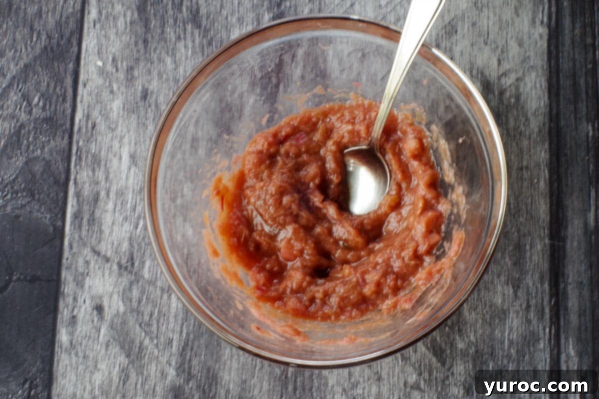 Smooth rhubarb purée in a clear glass bowl with a spoon, after brown sugar has been incorporated, enhancing its flavor.