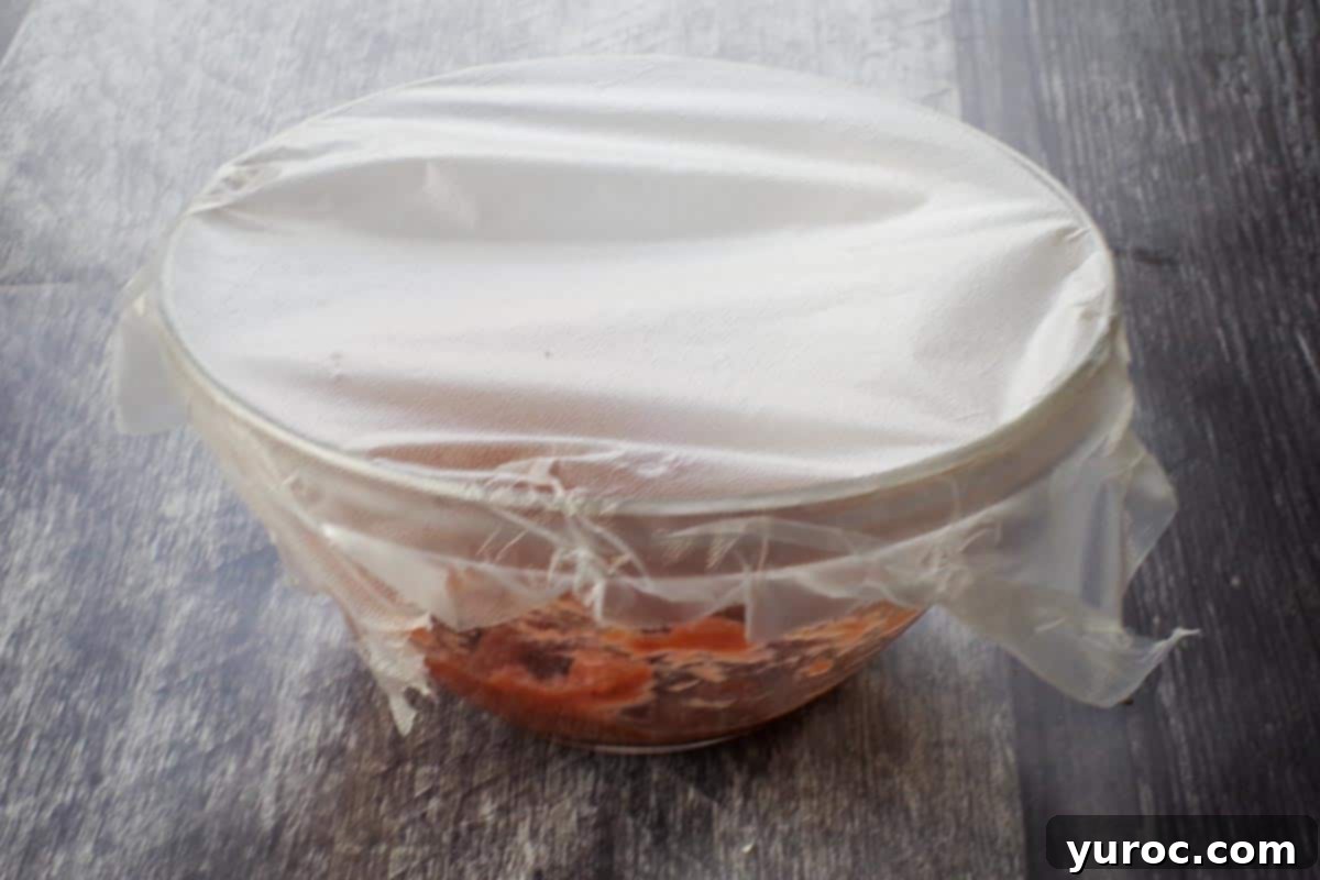 A bowl of sweetened rhubarb purée covered with plastic wrap, securely sealed for refrigeration.