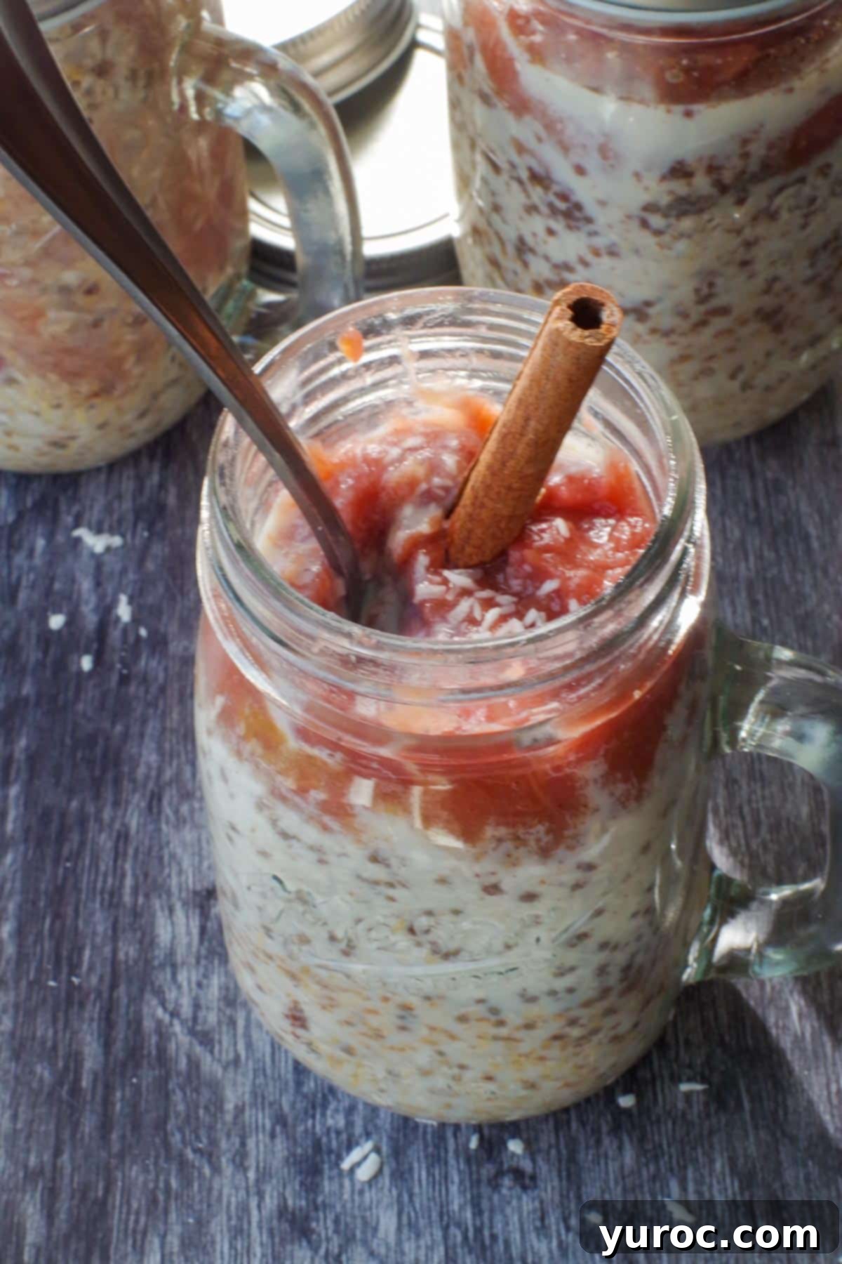 A jar of Rhubarb Overnight Oats with two more blurred jars in the background, showcasing the vibrant pink purée and a sprinkle of coconut.