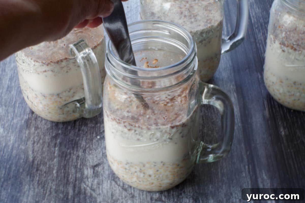 A spoon gently stirring the milk into the oat mixture within a glass mason jar mug, ensuring all ingredients are well combined.