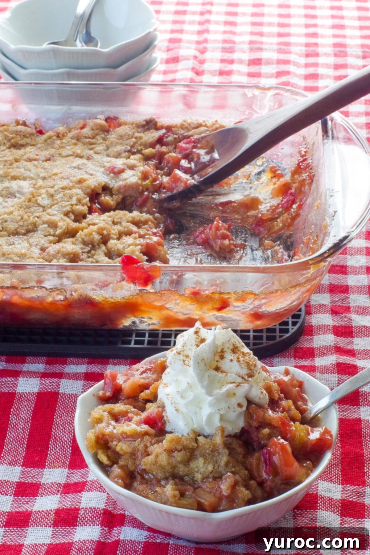 Delicious rhubarb crumble topped with a dollop of whipped cream, served in a charming white petal dish with a spoon. A glass baking pan of more rhubarb crumble is visible in the background, highlighting its rustic appeal.