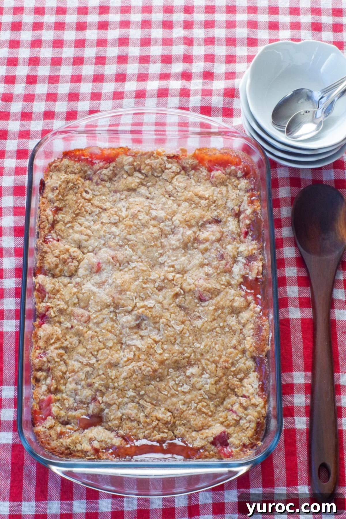 A beautiful, whole rhubarb crisp in a glass pan, perfectly baked and golden, adorned with a wooden spoon on one side and a stack of small white bowls and spoons on the other, hinting at serving.