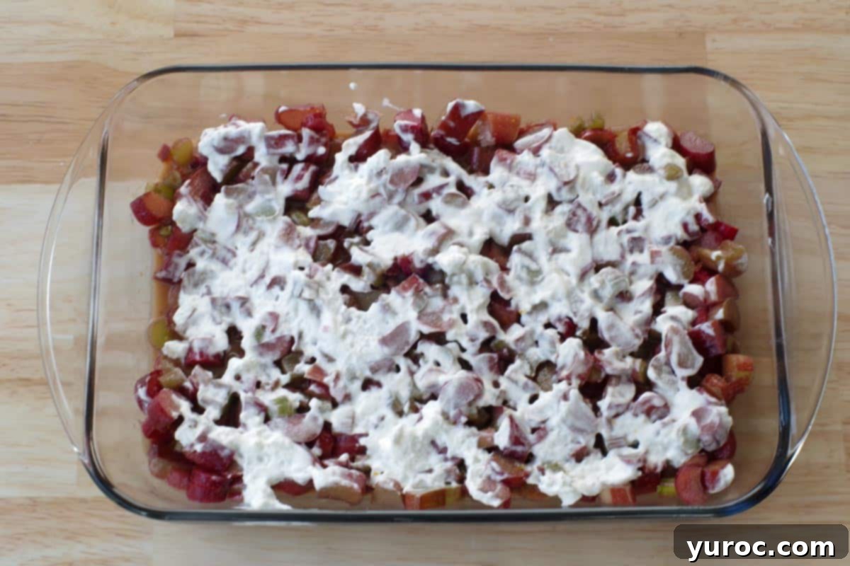 A thin, even layer of whipped cream delicately spread over the vibrant rhubarb mixture in the baking dish.