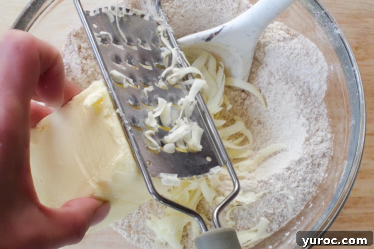 Cold butter being grated directly into the dry ingredients for the crumble topping in a glass bowl using a cheese grater, illustrating the 'Top Tip' for an even crumb.