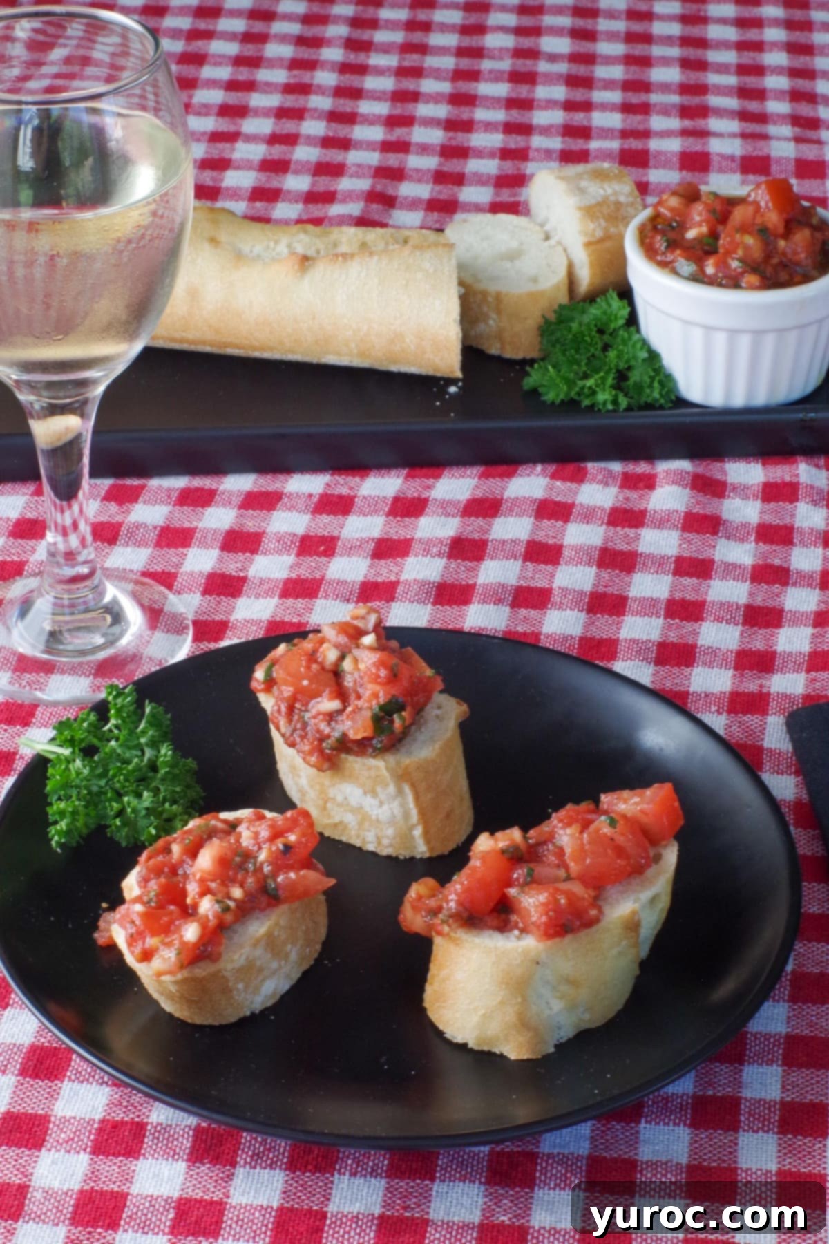 3 pieces of bread with bruschetta on a black plate with a baguette and bruschetta in the background