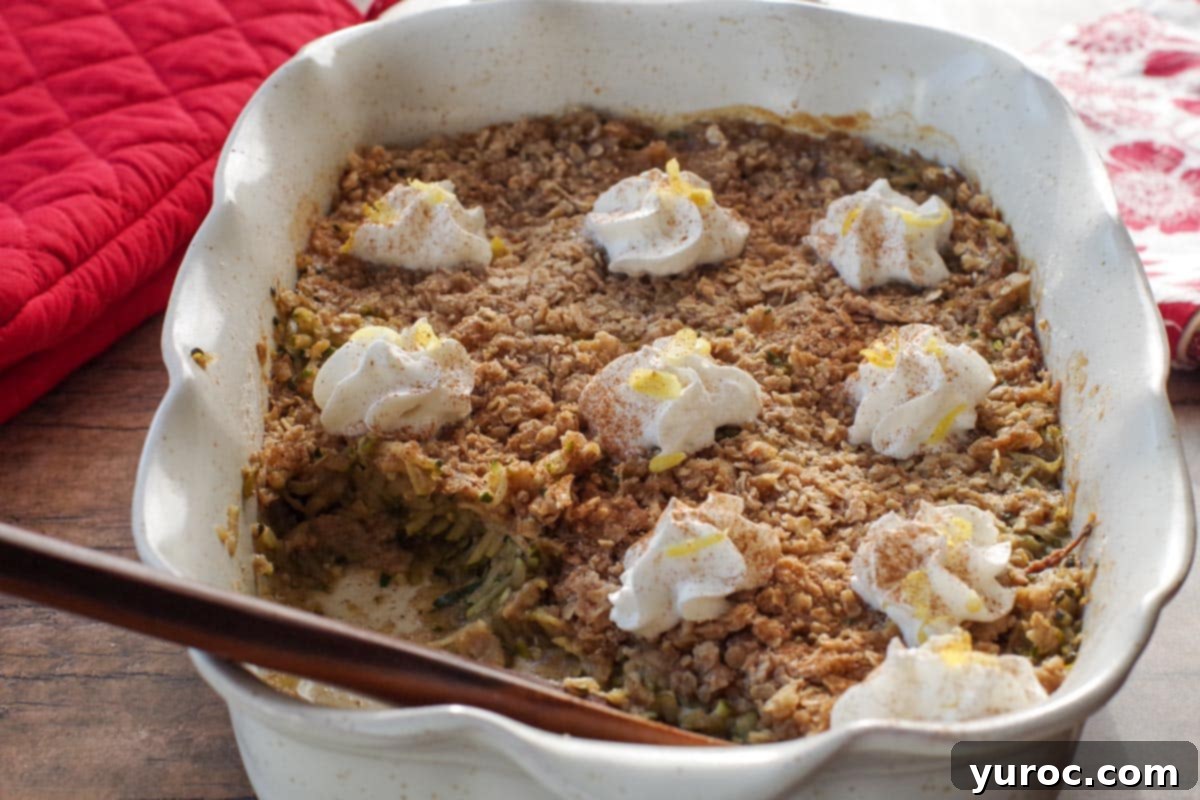 A large white baking dish filled with golden-brown zucchini crisp, topped with dollops of whipped cream. A wooden spoon rests in the dish, indicating a portion has been served, highlighting its irresistible appeal.