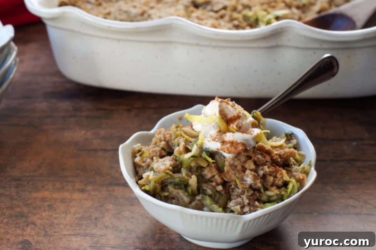A single serving of zucchini crisp in a small white bowl, with a larger baking dish of zucchini crisp visible in the soft-focus background, showcasing a freshly baked batch.