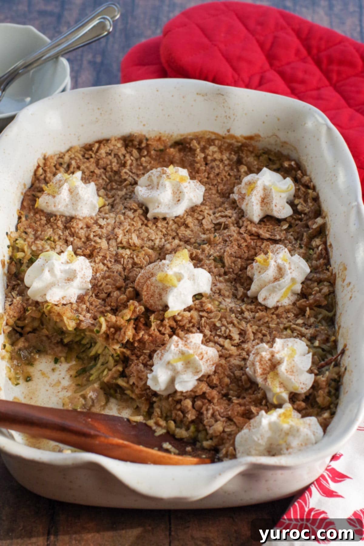 A large white baking dish filled with golden-brown zucchini crisp, topped with generous dollops of whipped cream. A wooden spoon is gently resting in the crisp, and a portion has been removed, highlighting its tempting texture.