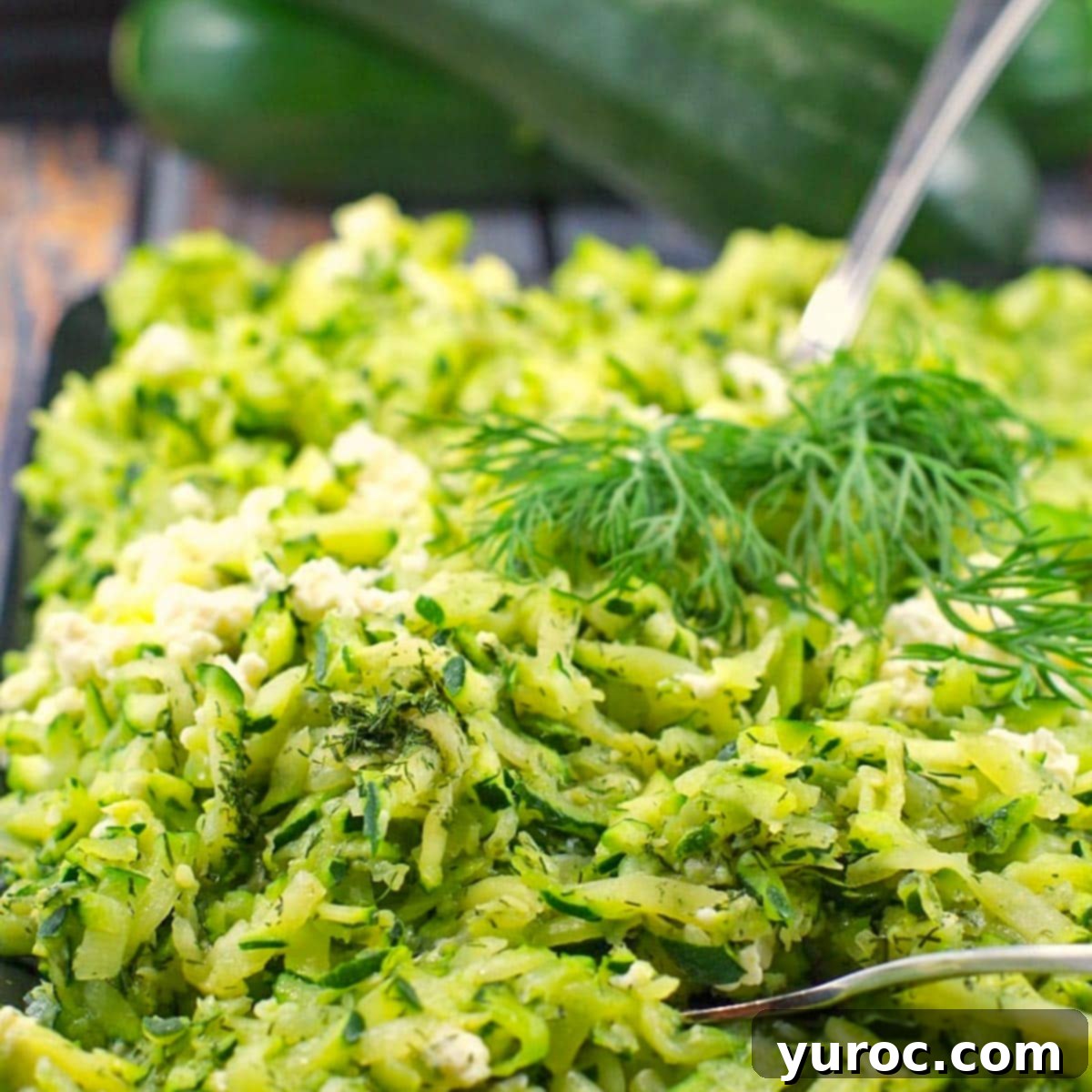 A close-up of finely shredded zucchini on a white plate, with whole zucchinis in the background, illustrating the ideal texture for recipes.