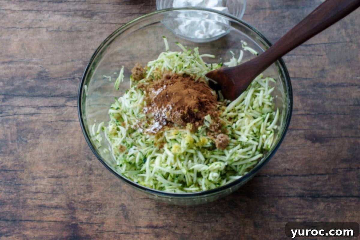 Freshly shredded zucchini in a large clear glass bowl, accompanied by a dark wooden spoon and other measuring ingredients, ready for mixing.