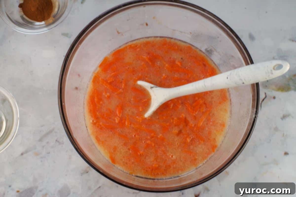 Glazed Carrot Cake Doughnut Muffins 7 Shredded carrots being gently folded into the wet ingredients mixture in a glass bowl.
