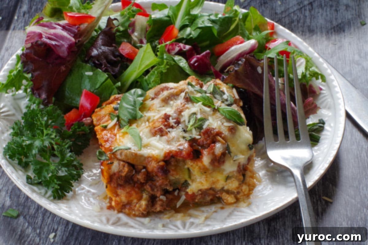 a slice of Zucchini Ground beef lasagna on a white plate with salad and a fork