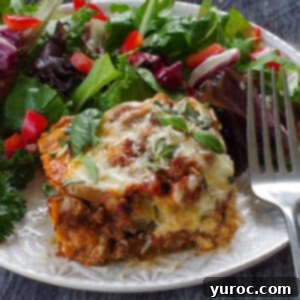 a slice of Zucchini Ground beef lasagna on a white plate with salad and a fork