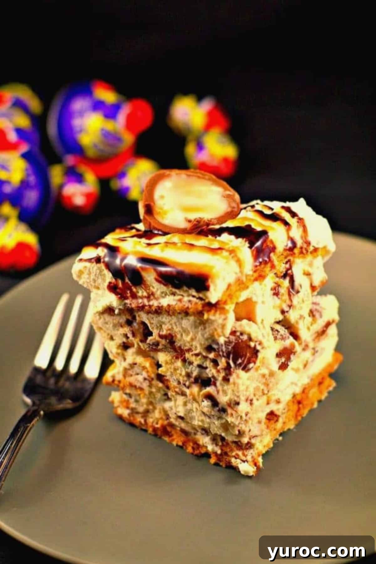 A slice of Easter Creme Egg Cake on a greenish grey plate with a fork, and wrapped Easter Creme Eggs in the background.