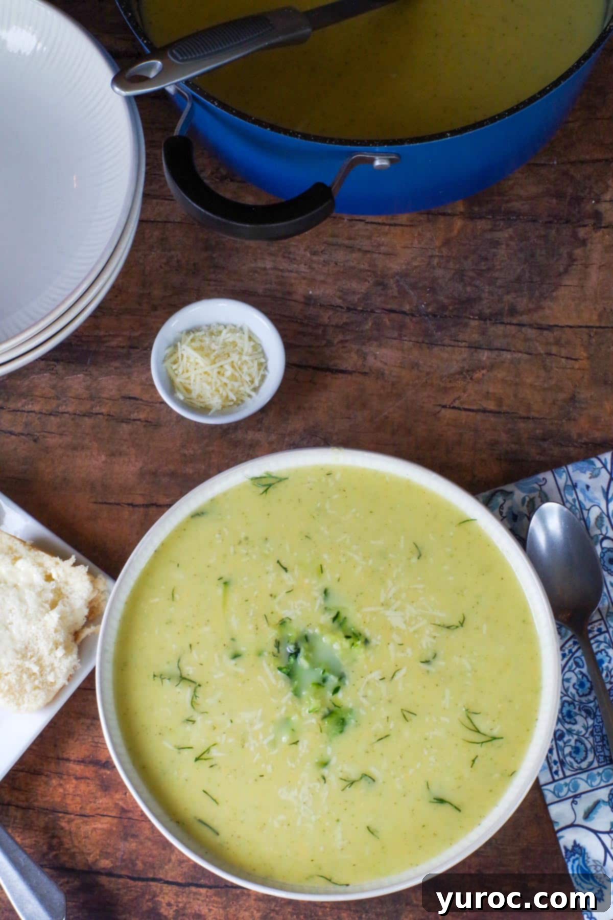 Wholesome Zucchini Potato Dill Soup 10 A bowl of creamy Zucchini Potato Soup in a beige bowl, served with a blue paisley napkin and a spoon, alongside a small white dish of grated Parmesan cheese in the background.