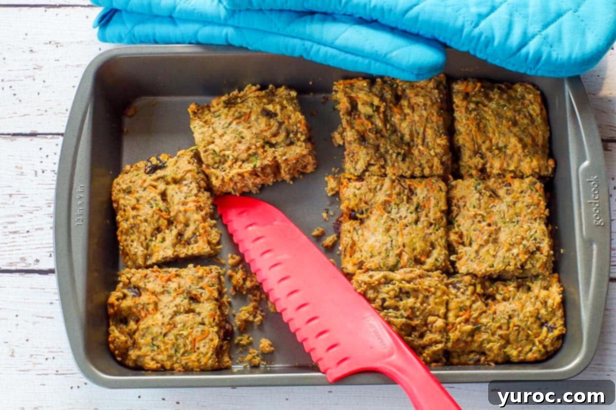 Freshly baked zucchini bars, perfectly cut in a pan, with a few slices already enjoyed, and a red silicone knife resting beside them.