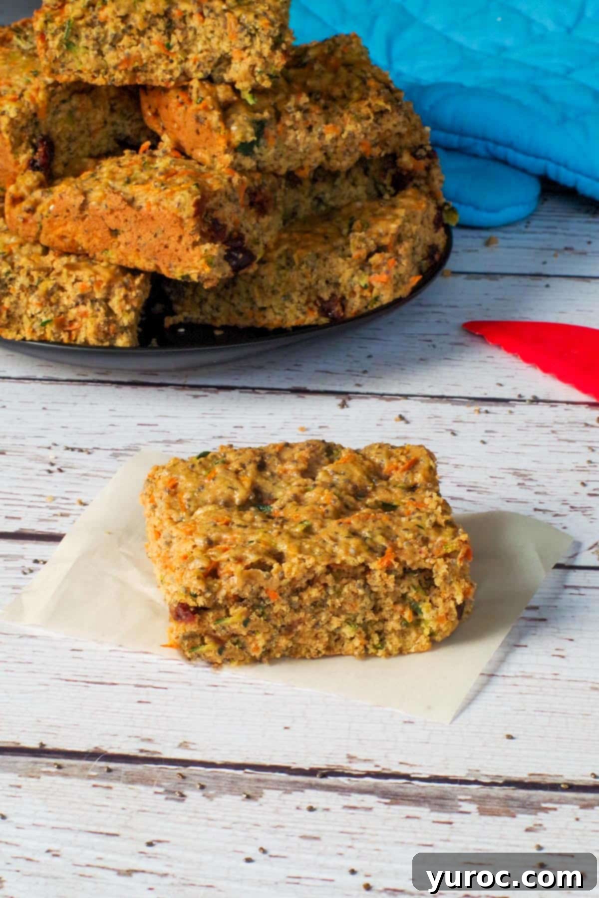 A healthy zucchini bar resting on a piece of parchment paper, with a rustic plate of more perfectly baked zucchini bars blurred in the background, showcasing its wholesome appeal.