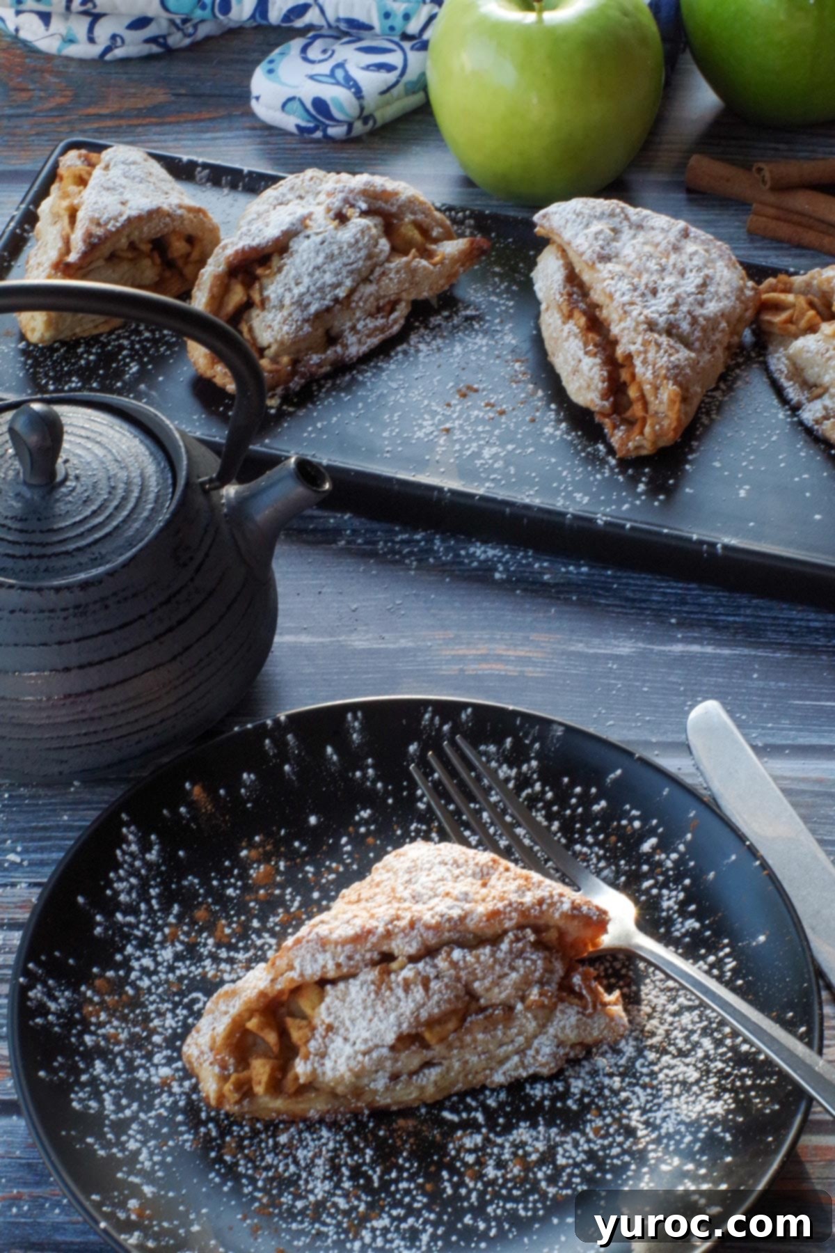 apple cinnamon scone on a black plate with a pot of tea, a tray of more scones and 2 green apples in the background