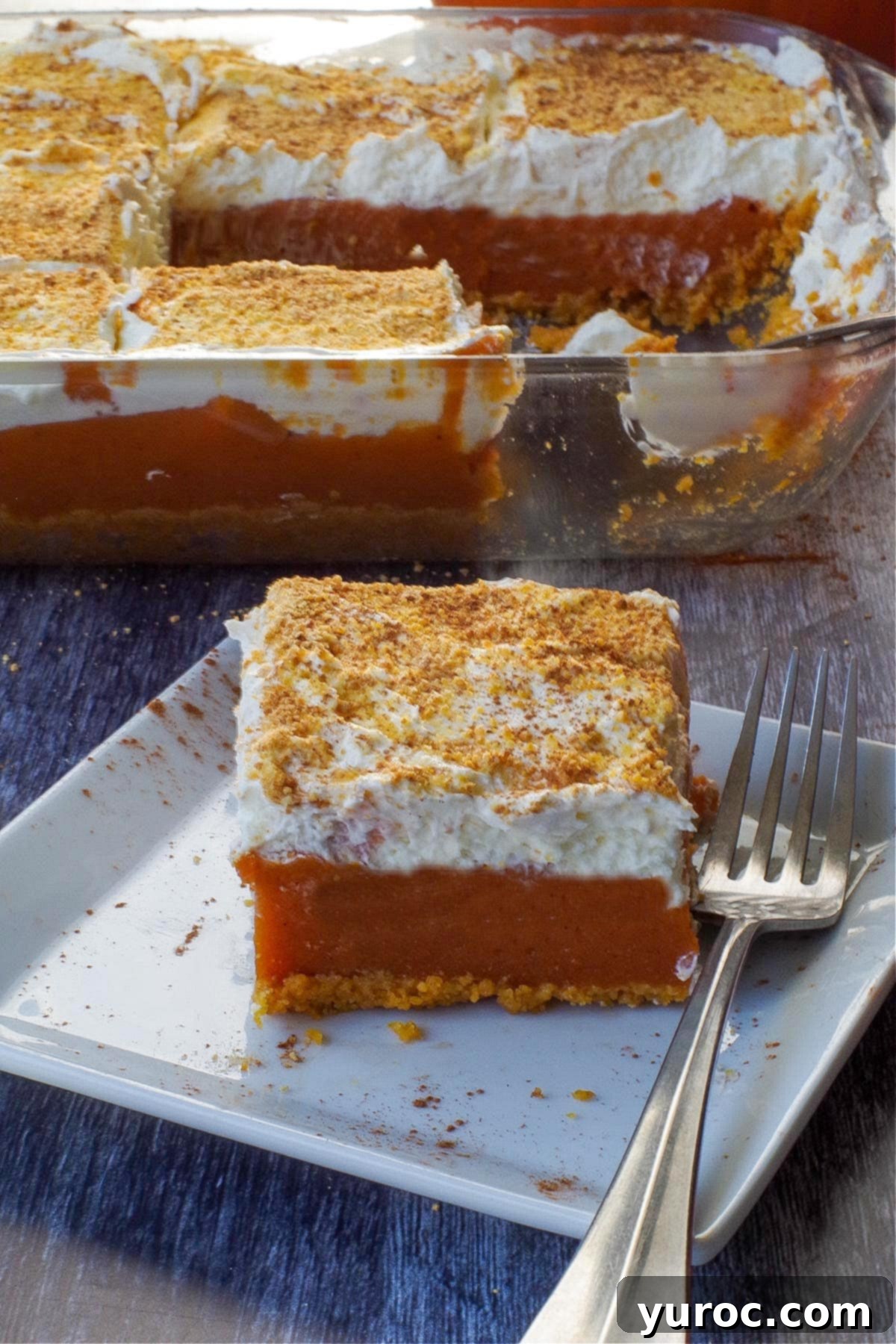 Effortless Pumpkin Perfection 17 A slice of layered pumpkin delight on a white plate with a fork, with the full pan of dessert in the blurred background, showcasing its perfect layers.