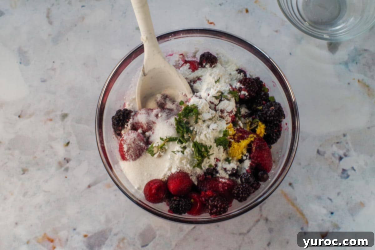 Berries with lemon, sugar, mint, and cornstarch in a glass bowl with a spoon.