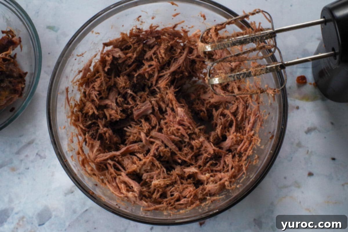 Pork being shredded in large glass bowl with electric hand mixer.