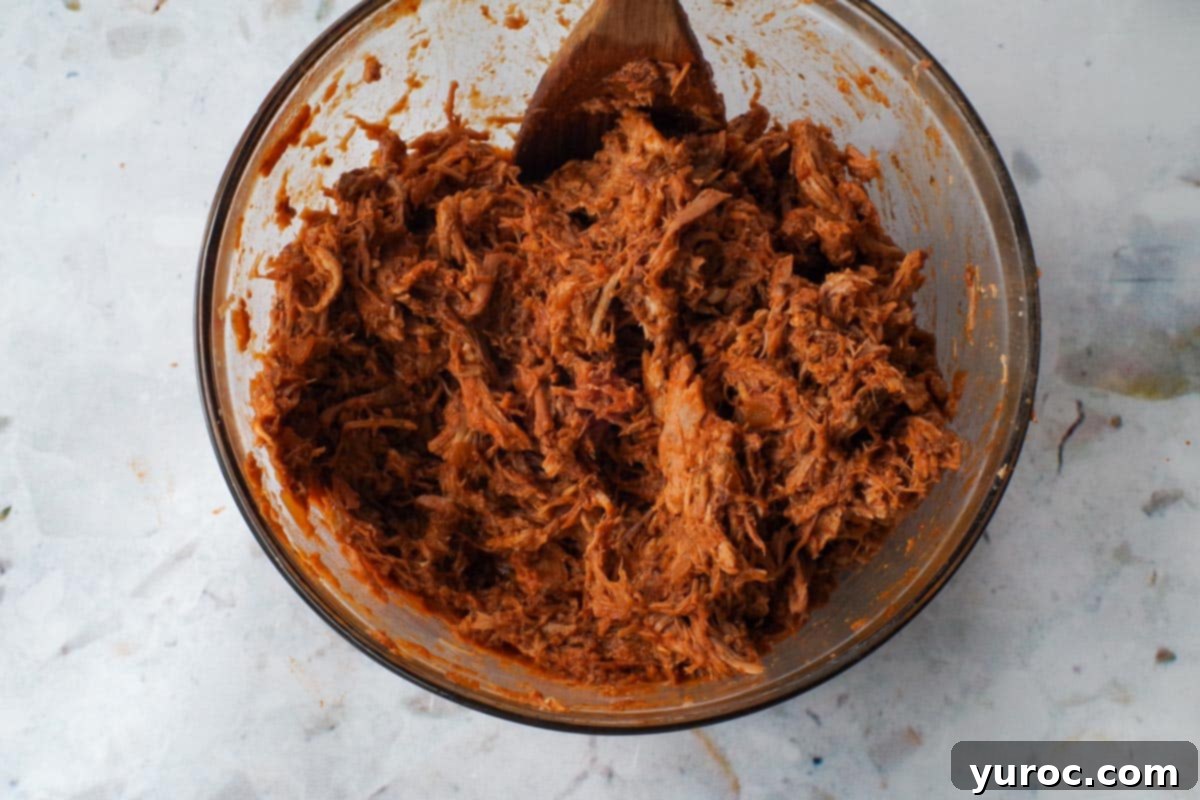 Reduced sauce added to shredded pork in a large glass bowl.