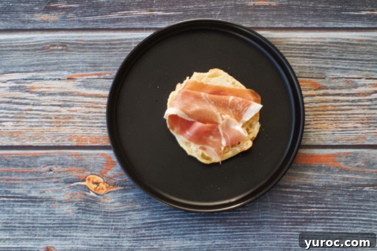 Prosciutto added to English muffin half on black plate.
