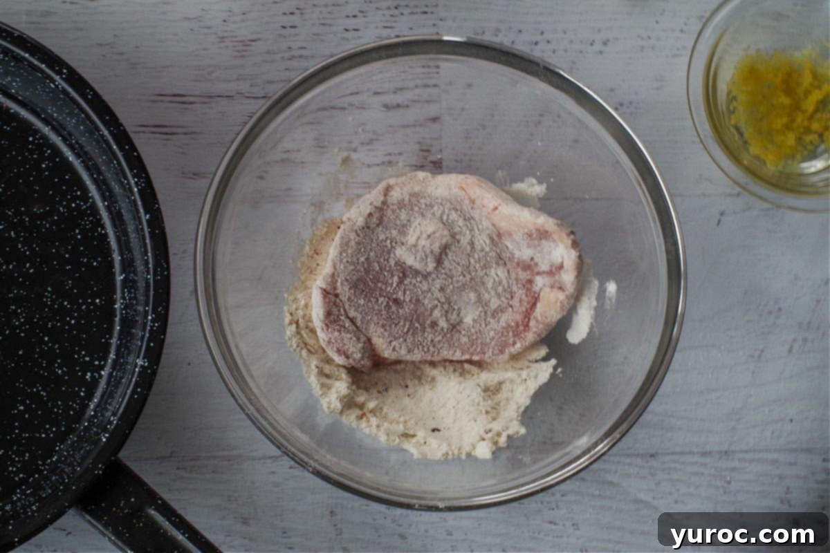 A piece of pork loin being coated with the flour mixture in a medium glass bowl.