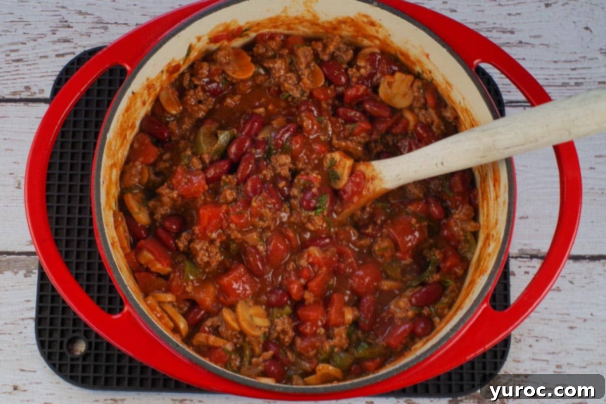 chili in orange dutch oven with wooden spoon with green peppers and onions cooked through