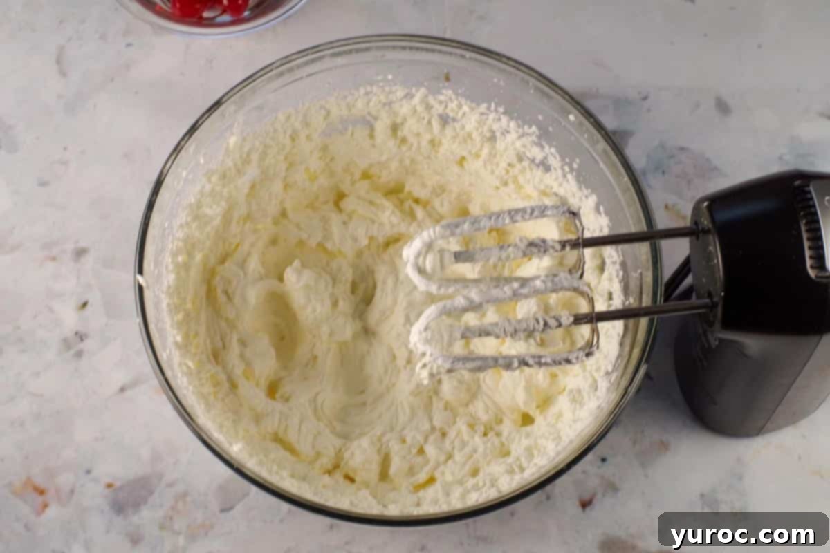 Whipping cream whipped to stiff peaks in a large glass bowl, with beaters from an electric mixer over the bowl.