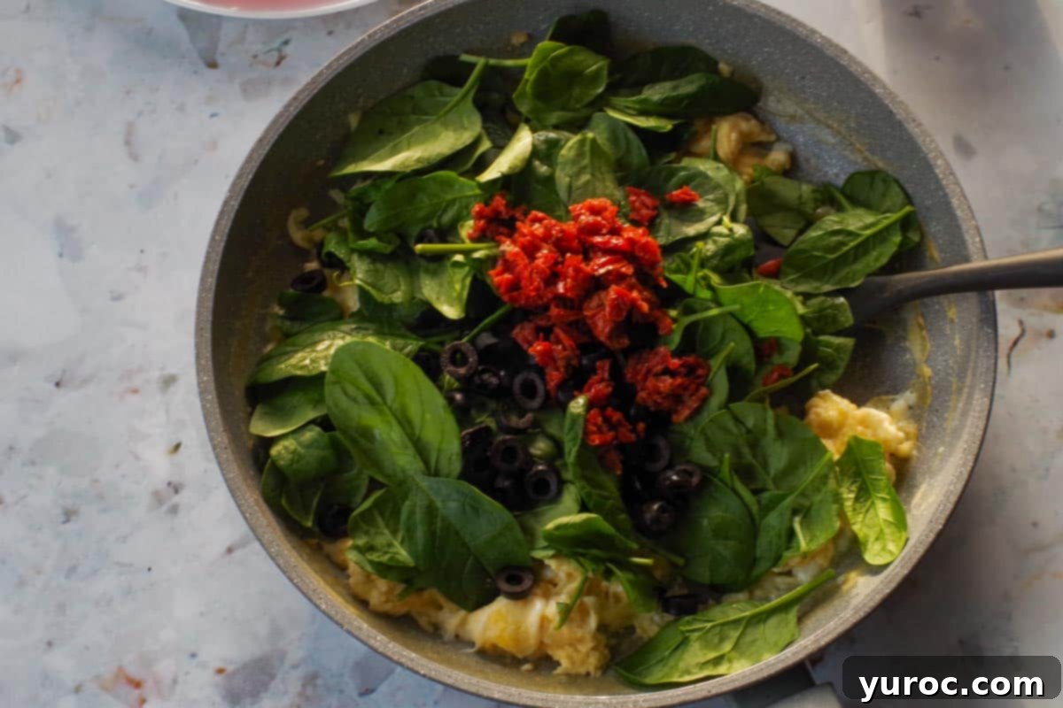 Spinach. olives and sun-dried tomatoes added to eggs in frying pan.