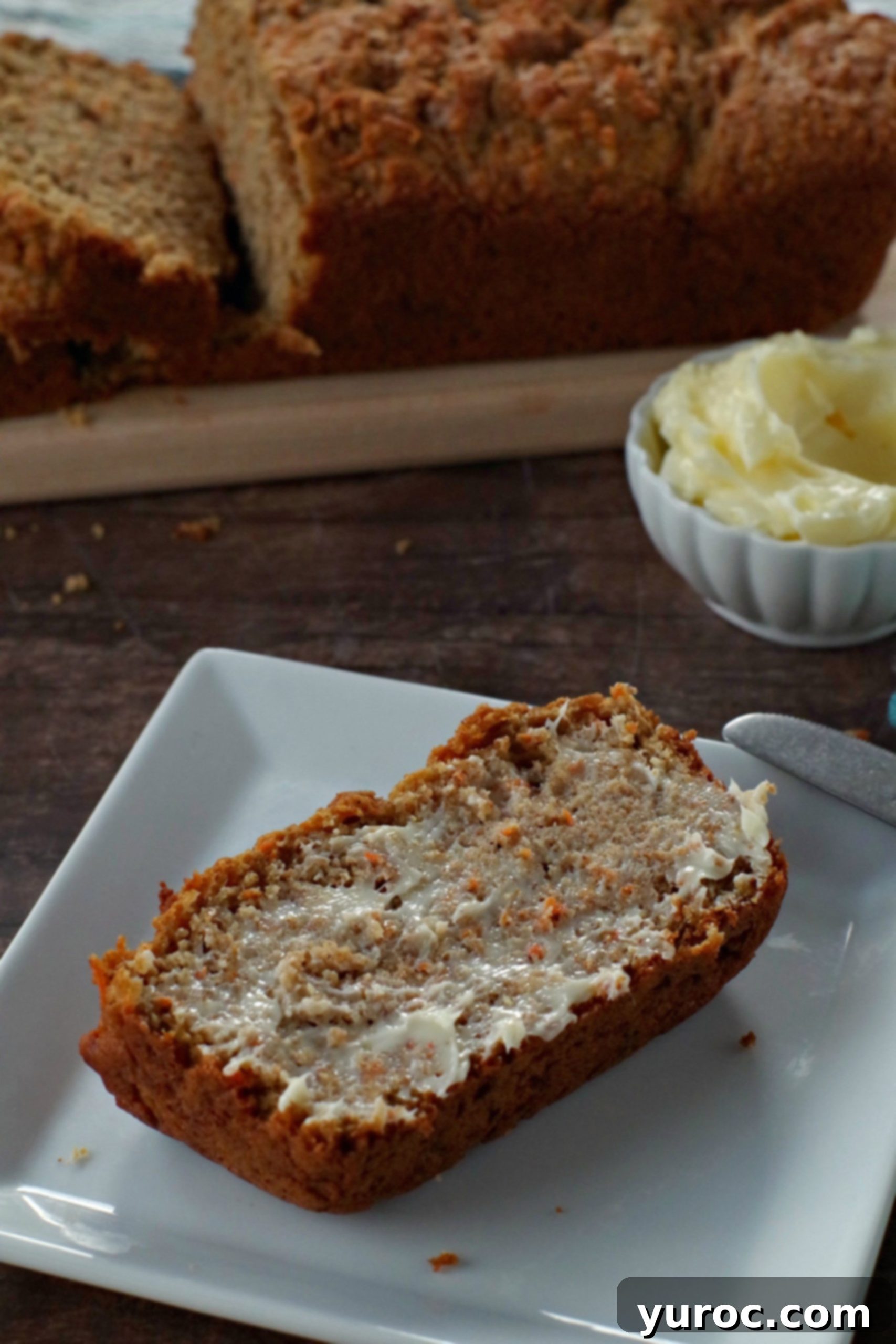 healthy carrot loaf slice with butter on a white plate with white dish of butter in the background. There is also a narrow wooden bread board in the background with the remainder of the carrot loaf on it (partially sliced)