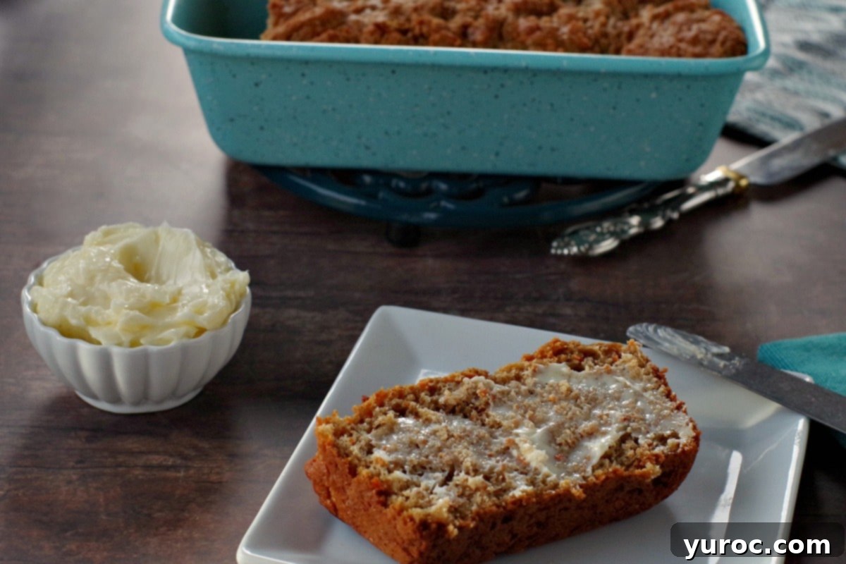 healthy carrot loaf slice with butter on a white plate with white dish of butter in the background. There is also a blue loaf pan in the background with the remainder of carrot loaf in it, as well as a silver knife beside the loaf pan.