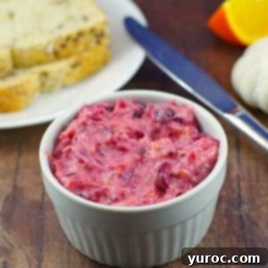 Creamy Cranberry Aioli in a white dish on a brown surface, with fresh bread, a knife, garlic, and orange slices in the background.