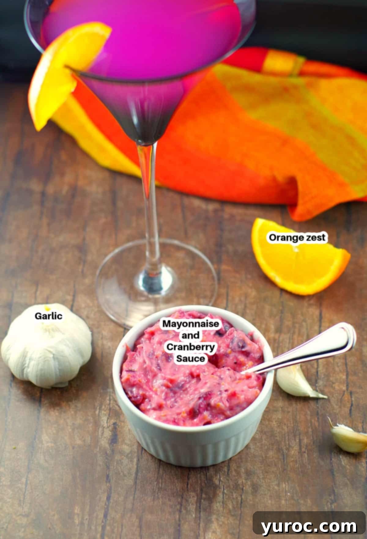 Cranberry aioli ingredients laid out on a wooden surface with a Cosmopolitan drink in the background, showcasing fresh cranberries, oranges, garlic, mayonnaise, and a dish of finished aioli.