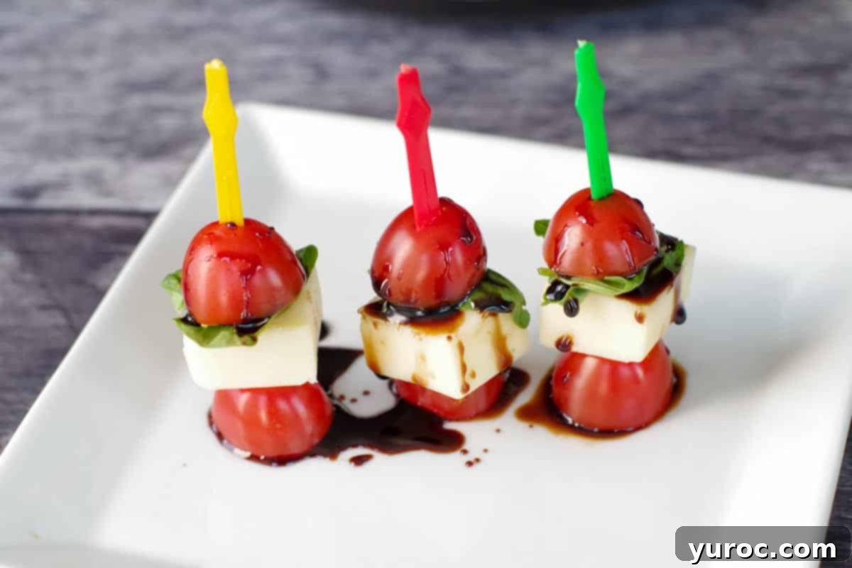 3 Caprese salad sticks on a white plate, drizzled with balsamic glaze