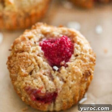 15 Deliciously Healthy Bran Muffin Recipes 9 A rustic Whole Wheat Raspberry Apple Bran Muffin resting on parchment paper, its top lightly dusted, with more delicious muffins blurred in the background on a wire rack, evoking homemade goodness and warmth.