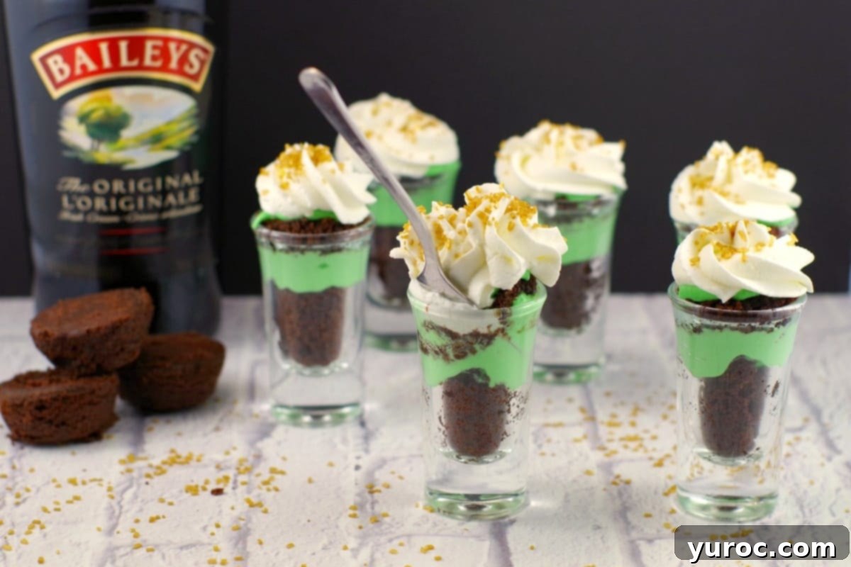 St. Patrick's Day Shamrock Irish Cream Shots 2 6 St. Patrick's Day Irish Cream Green Dessert Shots on a faux wood surface with a bottle of Baileys Irish Cream and 3 2-bite brownies in the background.