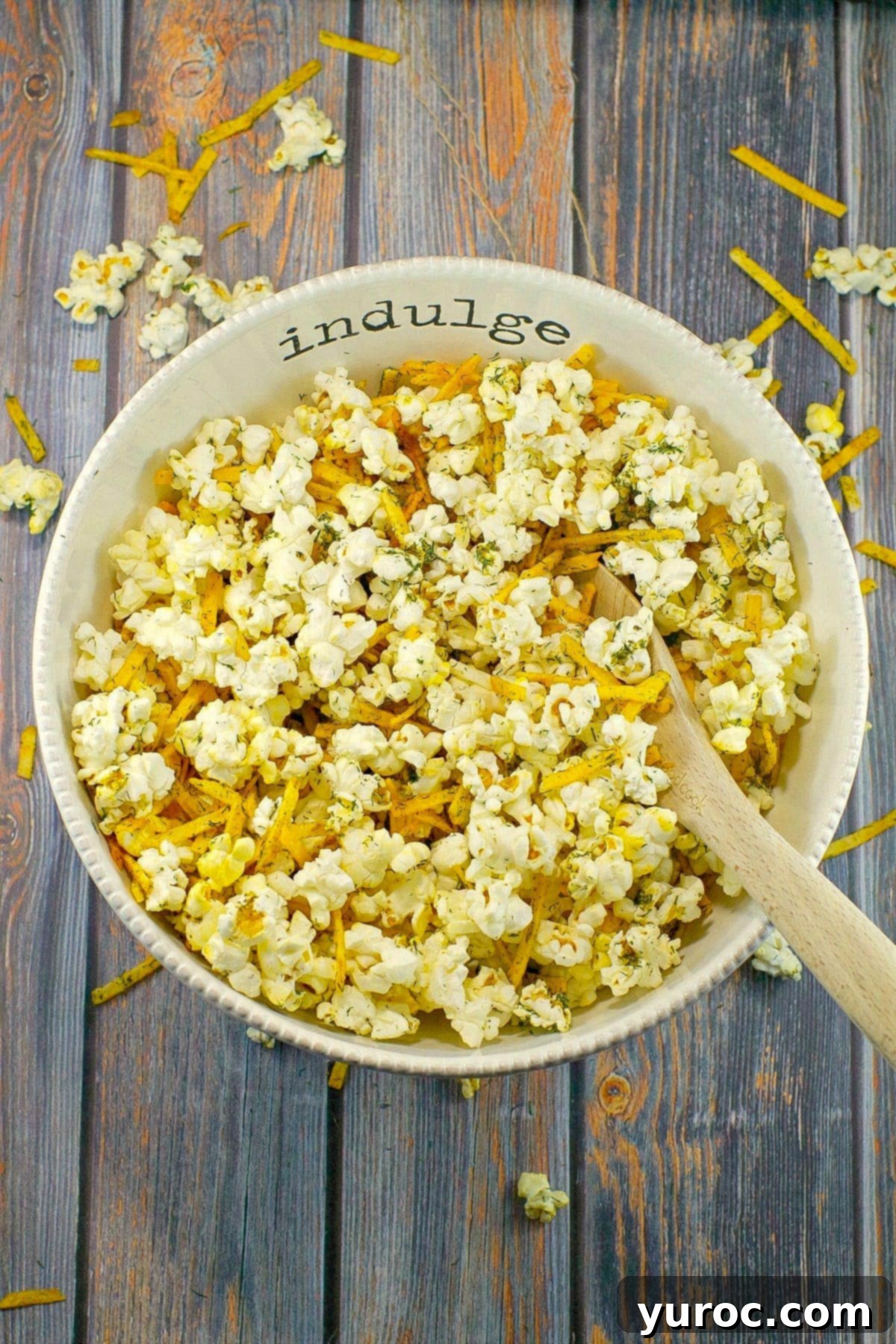 Savory popcorn in a white bowl, with a wooden spoon, with the words 'indulge' on it, on faux wooden surface.