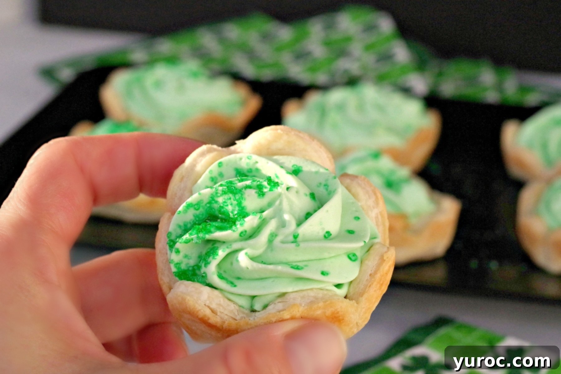 A vibrant green Shamrock tart being held up to showcase its creamy filling and delicate crust, with more tempting tarts artfully arranged on a tray in the background.