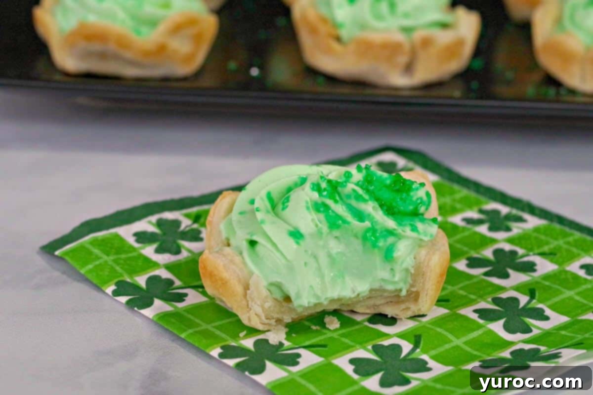 A delectable St. Patrick's Day Shamrock Tart sitting on a green shamrock patterned napkin, with a bite taken out revealing the creamy green filling. More whole tarts are artfully arranged on a serving tray in the softly blurred background.