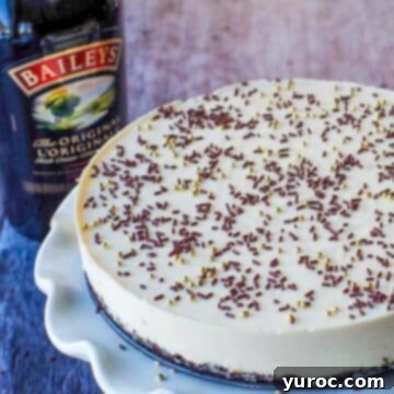 A tantalizing slice of a whole No-Bake Baileys cheesecake displayed on a white cake platter, with a bottle of Baileys in the soft background, ready for dessert.