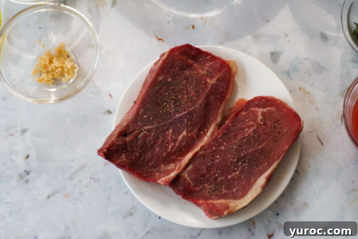 Two pieces of steak seasoned with salt and pepper on a white plate, ready for grilling.