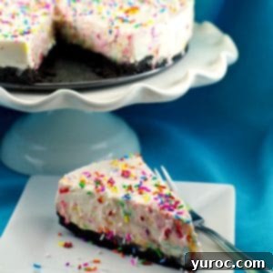 a slice of birthday cake cheesecake on a white plate with a fork on the side and the remainder of the cake on a white stand in the background